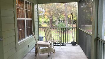 A covered porch with a ceiling fan, two chairs, a potted plant, and a view of a yard with trees and a fence.