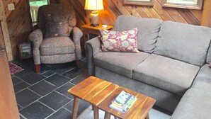 Ein gemütliches Zimmer mit einem gemusterten Sessel, einem grauen Sofa, einem Holztisch und einer Lampe. An der Wand befinden sich gerahmte Bilder und ein Fenster mit Blick auf Bäume.