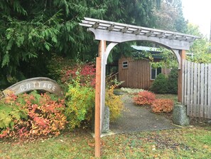 Een houten boogpoort die leidt naar een huisje met de tekst 'COTTAGE', een tuinpad en diverse struiken.