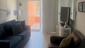 A living room with a dark blue sofa, a brown armchair, a ceiling fan, a television on a stand, and a sliding glass door leading to an outdoor area. - Coquina Beach Club 110, Beachfront Studio. Booking late summer weeks. (Bradenton Beach)