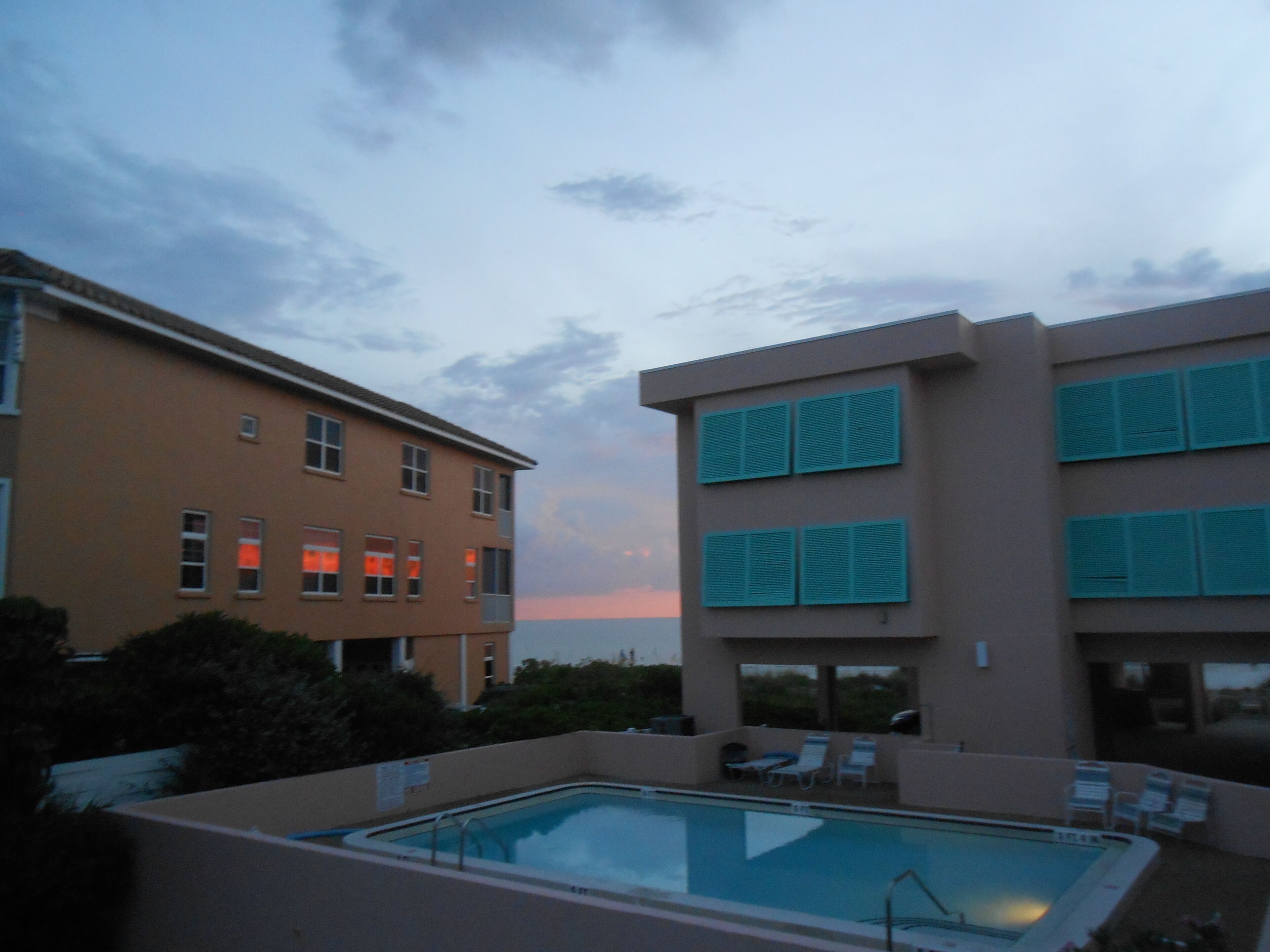 A swimming pool with lounge chairs, a building with multiple windows, and a sunset in the background.