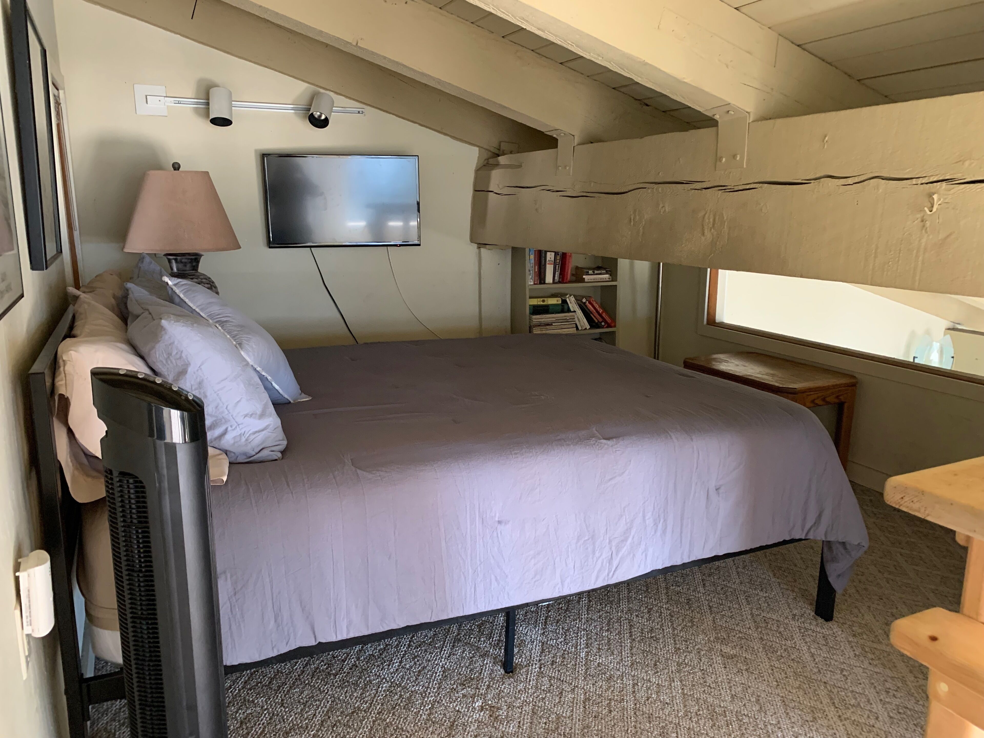 Ein Schlafzimmer mit einem Bett, einem an der Wand befestigten Fernseher, einer Lampe, einem Bücherregal und einem Fenster.