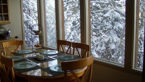 A dining area with a round table set for four, surrounded by wooden chairs. A window provides a view of snow-covered trees outside. - LUXURY VIEW CONDO/SKI-IN/SKI-OUT/TO CANYON/EAGLE EXPRESS (Mammoth Lakes)