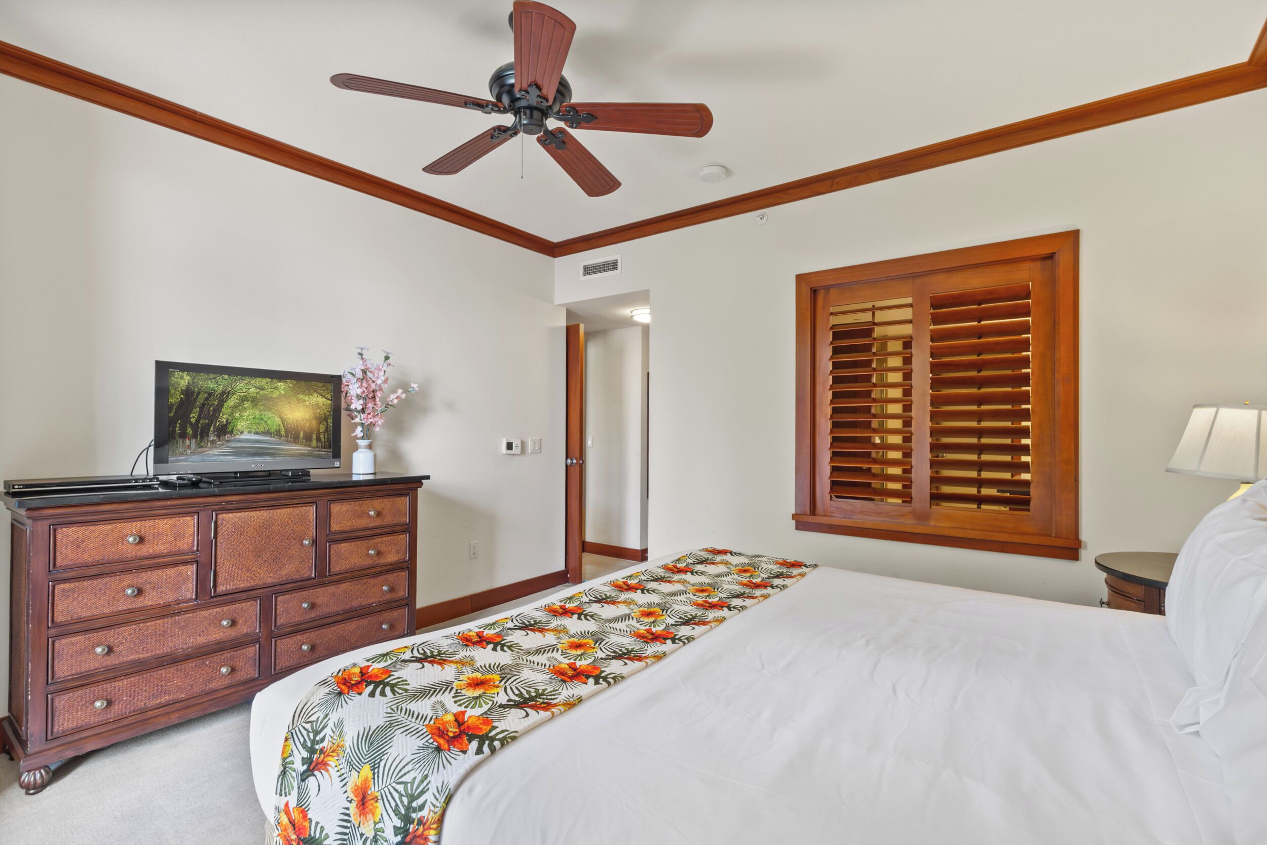 Een slaapkamer met een bed, een houten dressoir, een plafondventilator, een televisie op het dressoir, en een raam met houten luiken.