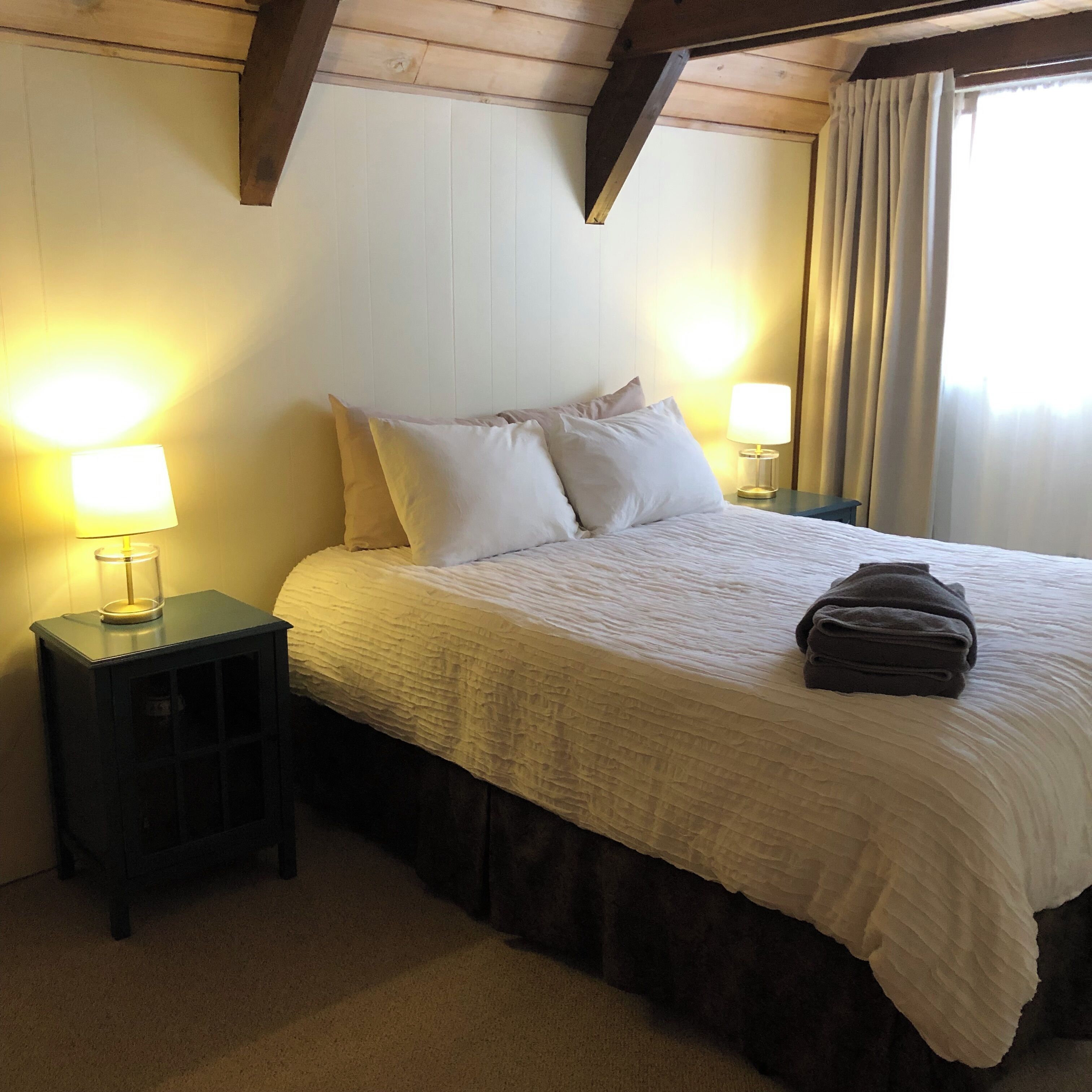 A neatly made bed with white bedding, two bedside lamps, and a window with curtains.