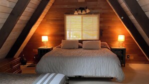 Ein gemütliches Schlafzimmer im Dachgeschoss mit einem großen Bett, zwei Nachttischen mit Lampen und einem Fenster mit Jalousien.