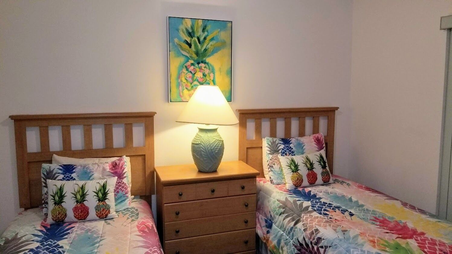 A bedroom with two single beds, each with a pineapple-themed pillow, a wooden nightstand with a lamp, and a wall painting of a pineapple.