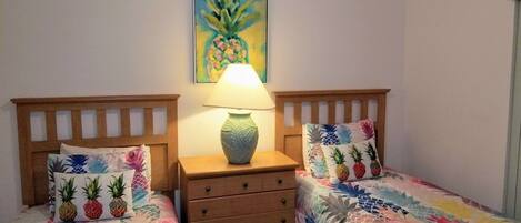 A bedroom with two single beds, each with a pineapple-themed pillow, a wooden nightstand with a lamp, and a wall painting of a pineapple.