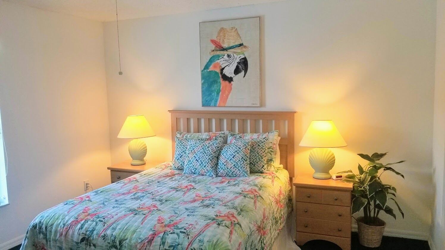 A bedroom with a bed, two bedside lamps, a nightstand, a potted plant, and a wall painting.