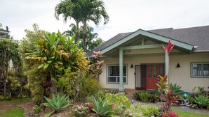 Een huis met een veranda, omringd door een tuin met verschillende planten en een palmboom.