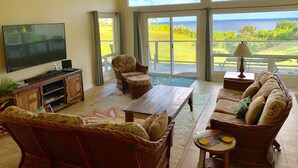 Een ruime woonkamer met een grote flatscreen-tv, comfortabele zitplaatsen en uitzicht op een weelderig landschap door grote schuifdeuren.