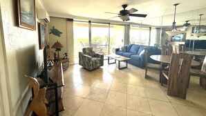 A spacious living area with a blue sofa, a coffee table, and a dining table with chairs. There is a ceiling fan, large windows, and a view of the outdoors. - On the beach, Beautiful ocean views. (Kihei)