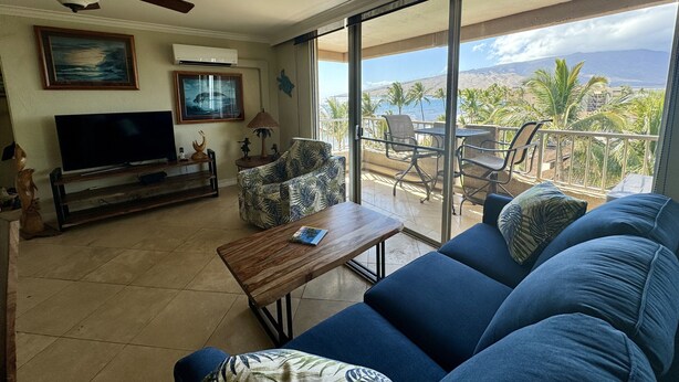 A living room with a sofa, a coffee table, and a TV. There is a balcony with a view of the ocean and palm trees. - On the beach, Beautiful ocean views. (Kihei)