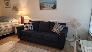 A living room with a dark sofa, a lamp, a painting of a beach, and a fan. - Come for the beach, stay for the memories. Enjoy free beach access & huge pool! (Atlantic Beach)