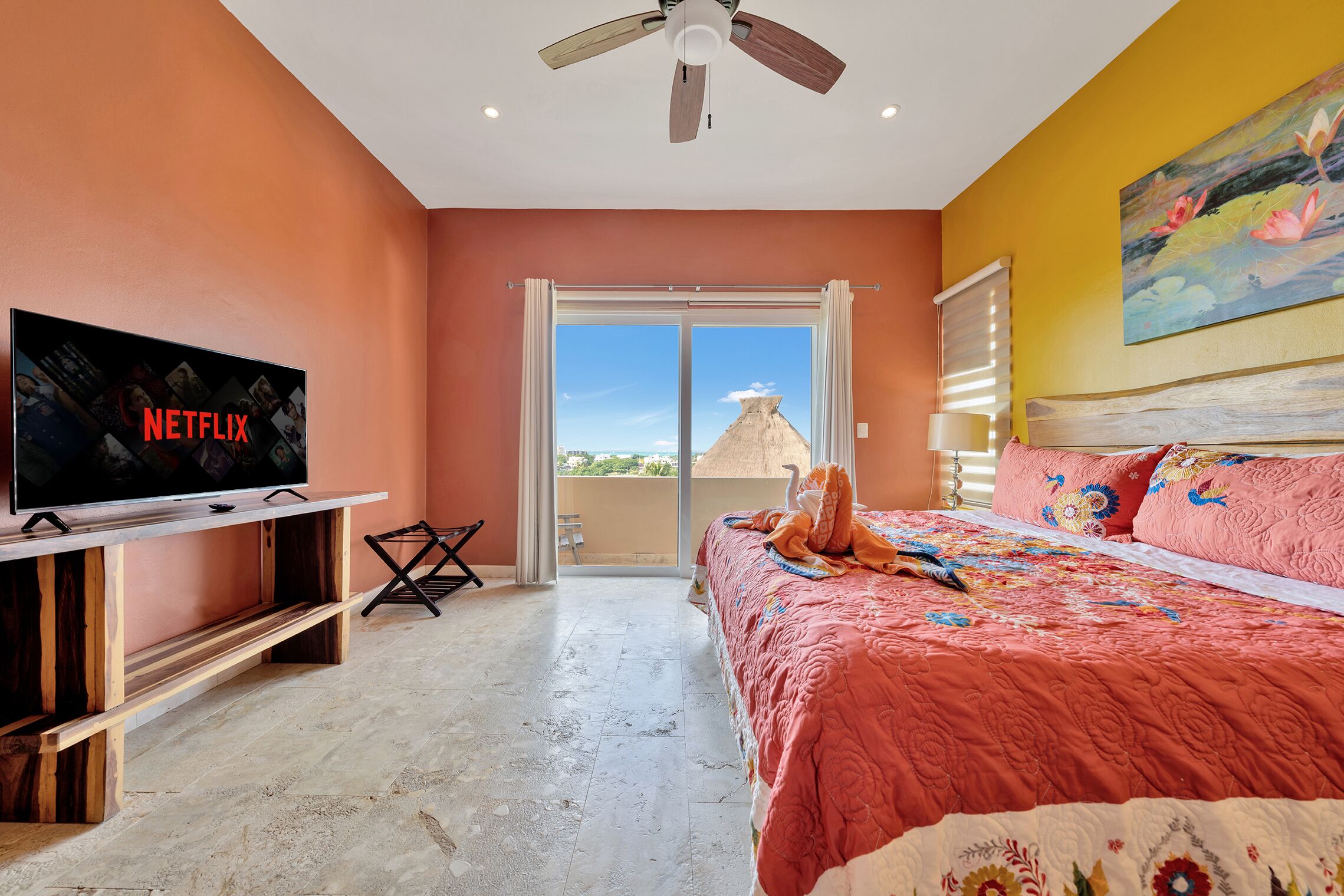 A bedroom with a bed, a TV on a wooden stand, a ceiling fan, and a view of the outside.