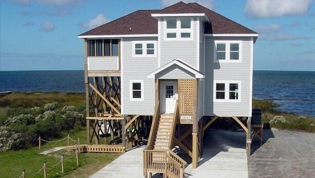 Ein Strandhaus mit zwei Etagen, einer Holzterrasse und einer Treppe, die zum oberen Stockwerk führt.