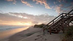 Eine Holztreppe führt zu einem Sandstrand mit Fußabdrücken, der sich unter einem Sonnenuntergangshimmel erstreckt.