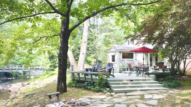 Un chalet rustique avec un toit rouge, un foyer en pierre et une terrasse en bois donnant sur une rivière.