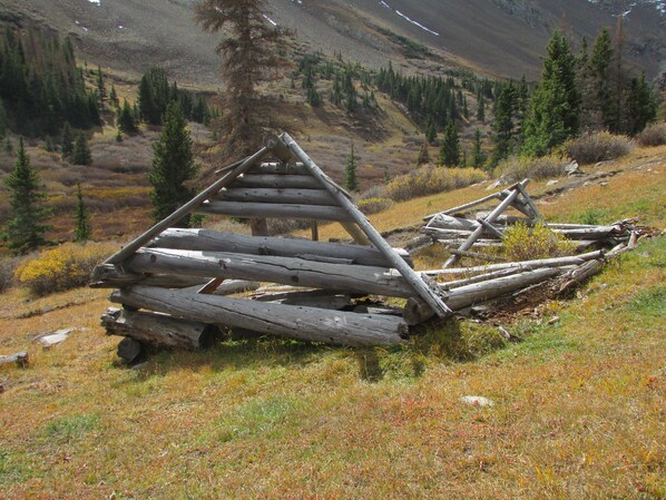 A dilapidated wooden structure in a mountainous area with sparse vegetation and a backdrop of trees and hills. - Sit on Your Deck in an Alpine Forest & Next to Mountain Stream (Westcliffe)
