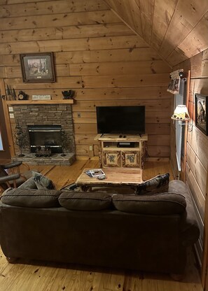 A cozy cabin-style living room with a stone fireplace, a television on a wooden stand, and a leather sofa.