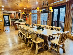 A wooden interior with a large dining table and chairs, a kitchen area, and large windows.