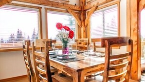 Une table à manger en bois, accompagnée de six chaises, un vase de fleurs et de grandes fenêtres donnant sur un paysage arboré.