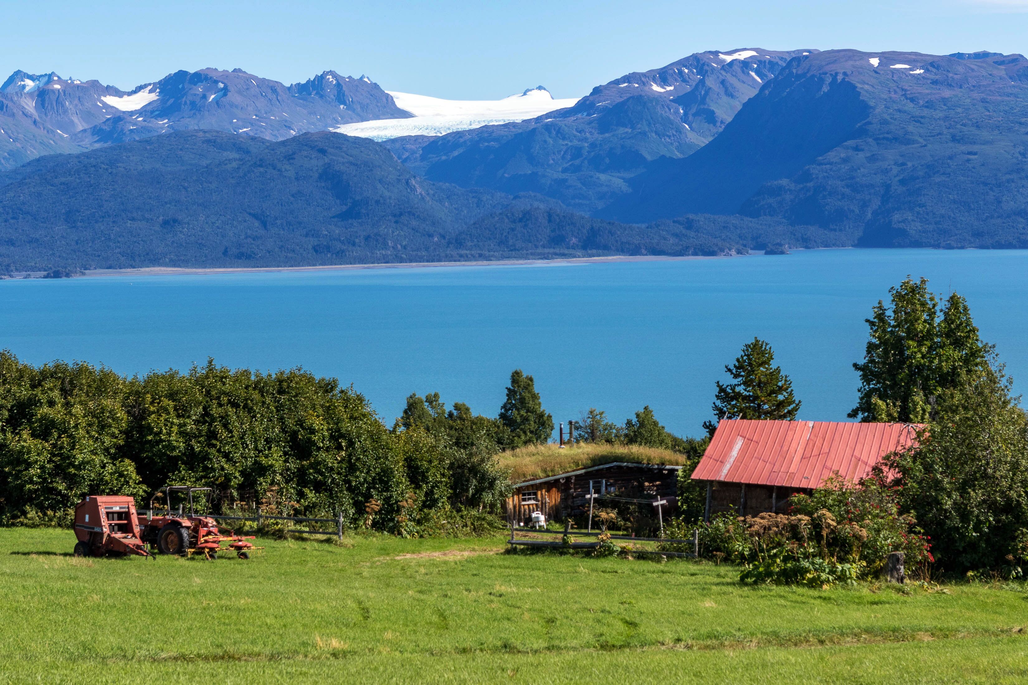 Un paysage rural avec une maison, un tracteur et un lac, les montagnes se dressant à l’arrière-plan.