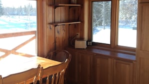 Interior de una cabaña de madera con una mesa de comedor, sillas, una ventana que ofrece vistas de un paisaje nevado y una estantería con un tostador.