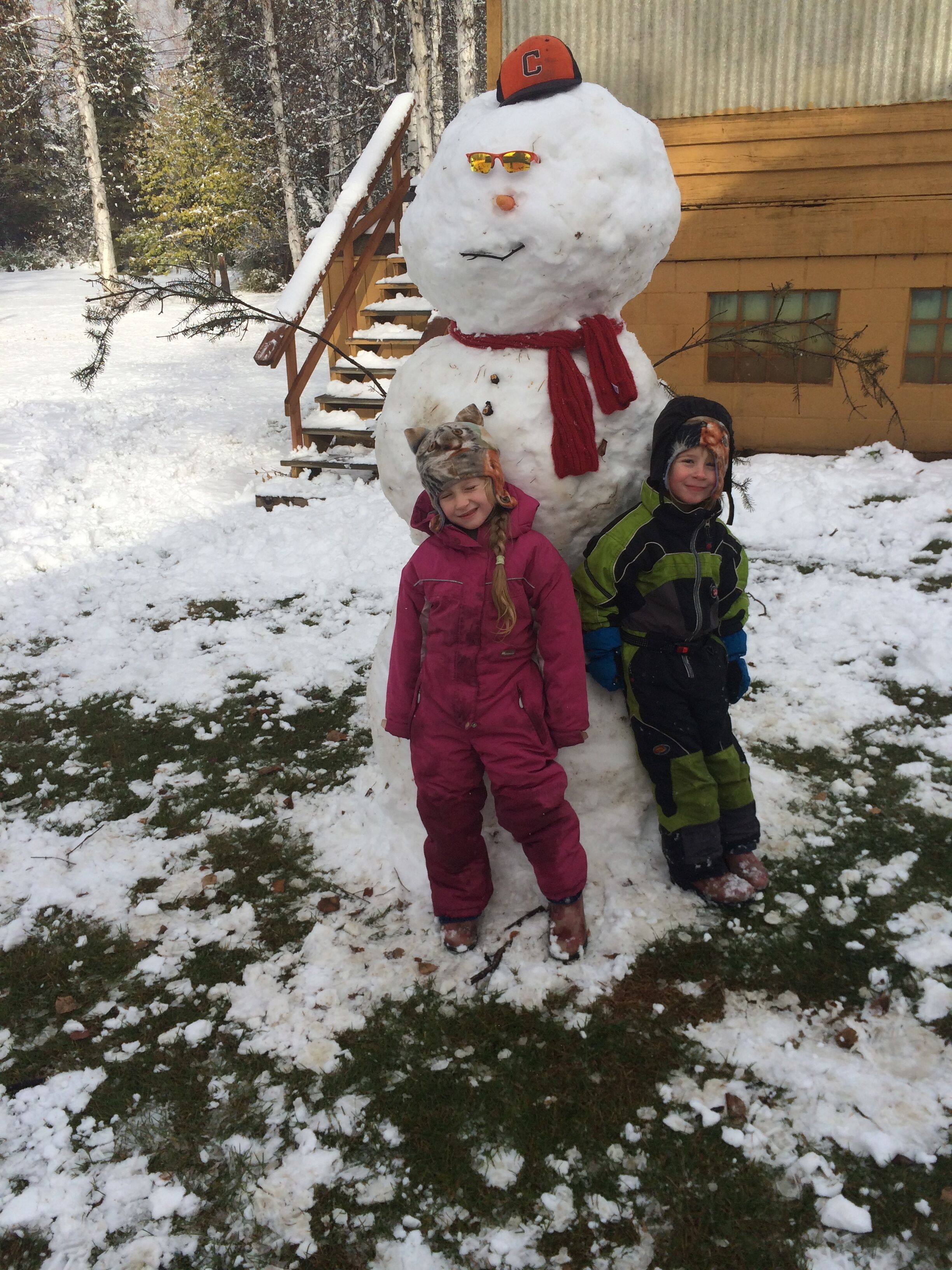 Zwei Kinder stehen vor einem Schneemann mit rotem Schal und Hut.