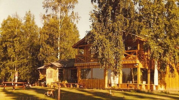 Una casa de madera rústica con balcón, rodeada de árboles y un patio cercado.