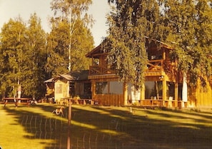 Una casa de madera rústica con balcón, rodeada de árboles y un patio cercado.