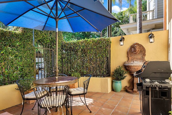 An outdoor patio with a round table, chairs, a blue umbrella, a grill, and a decorative wall.