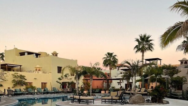 A swimming pool with lounge chairs and palm trees, surrounded by buildings with Mediterranean architecture.