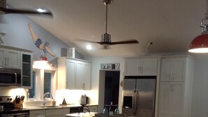Une cuisine moderne avec des meubles blancs, un îlot central doté de tabourets, un réfrigérateur et un ventilateur de plafond.