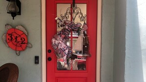 Une porte rouge ornée d’une guirlande décorative, un panneau en forme de tortue avec des chiffres, un paillasson et une petite plante en pot.