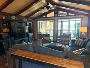 A spacious living room with a large television, comfortable seating, and a view through the glass doors.