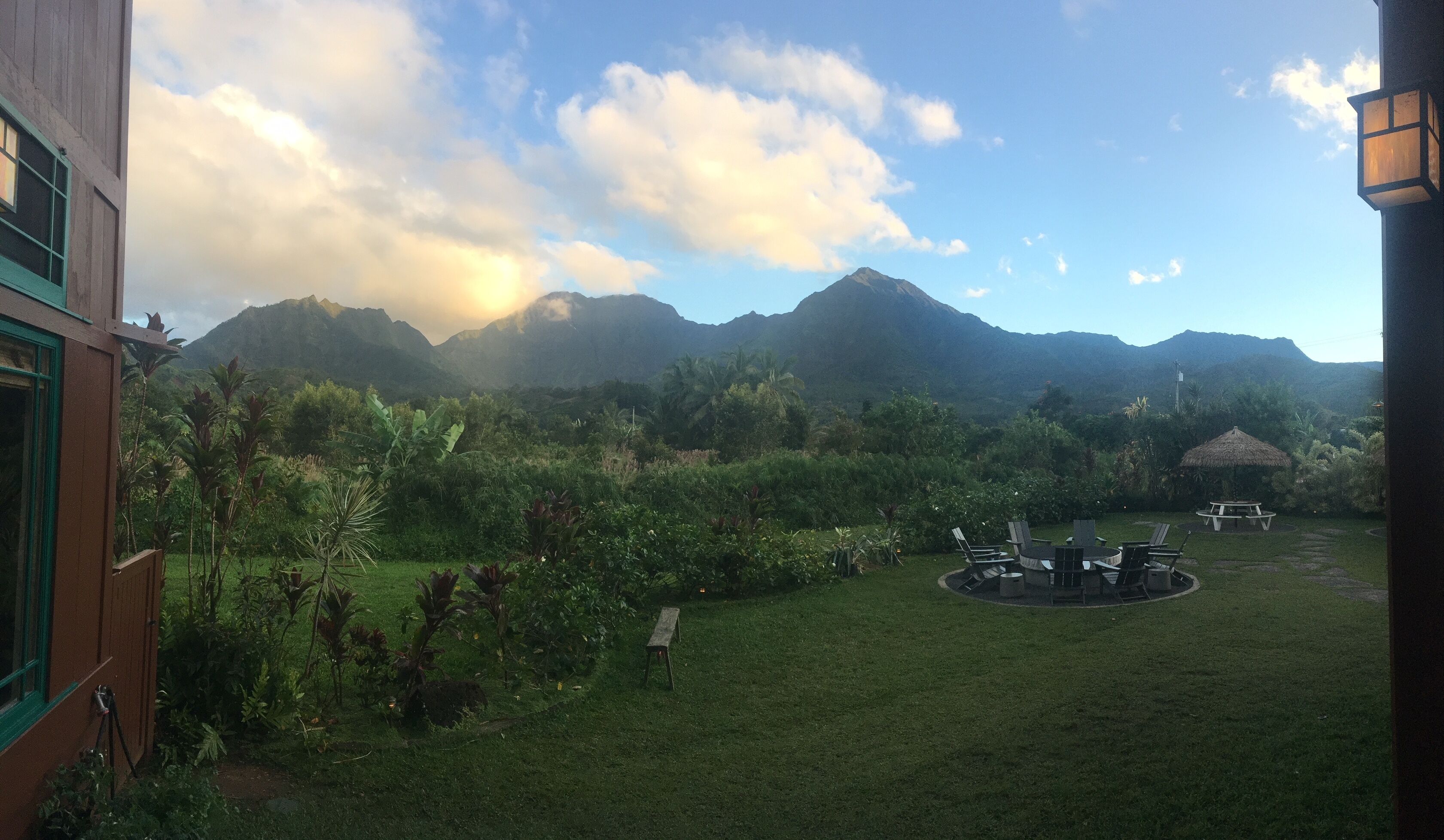 Une vue d’un jardin luxuriant avec un belvédère à toit de chaume, une table de pique-nique et des chaises, le tout entouré de plantes tropicales et avec une chaîne de montagnes en arrière-plan.
