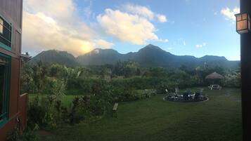 Une vue d’un jardin luxuriant avec un belvédère à toit de chaume, une table de pique-nique et des chaises, le tout entouré de plantes tropicales et avec une chaîne de montagnes en arrière-plan.