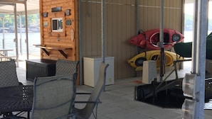 A covered outdoor seating area with metal chairs and a table, a wooden wall with a window, and kayaks stored against the wall. - 2 Master Suites, Incredible Views Sleeps 10-12, Great Dock (Sunrise Beach)