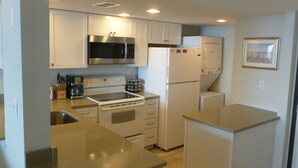 Una cocina compacta con armarios blancos, un microondas de acero inoxidable, un horno integrado, un refrigerador y una cafetera.