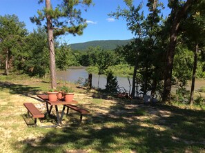 Ein Picknicktisch mit Bänken und Topfpflanzen in einer bewaldeten Gegend, im Hintergrund ein See und Hügel.
