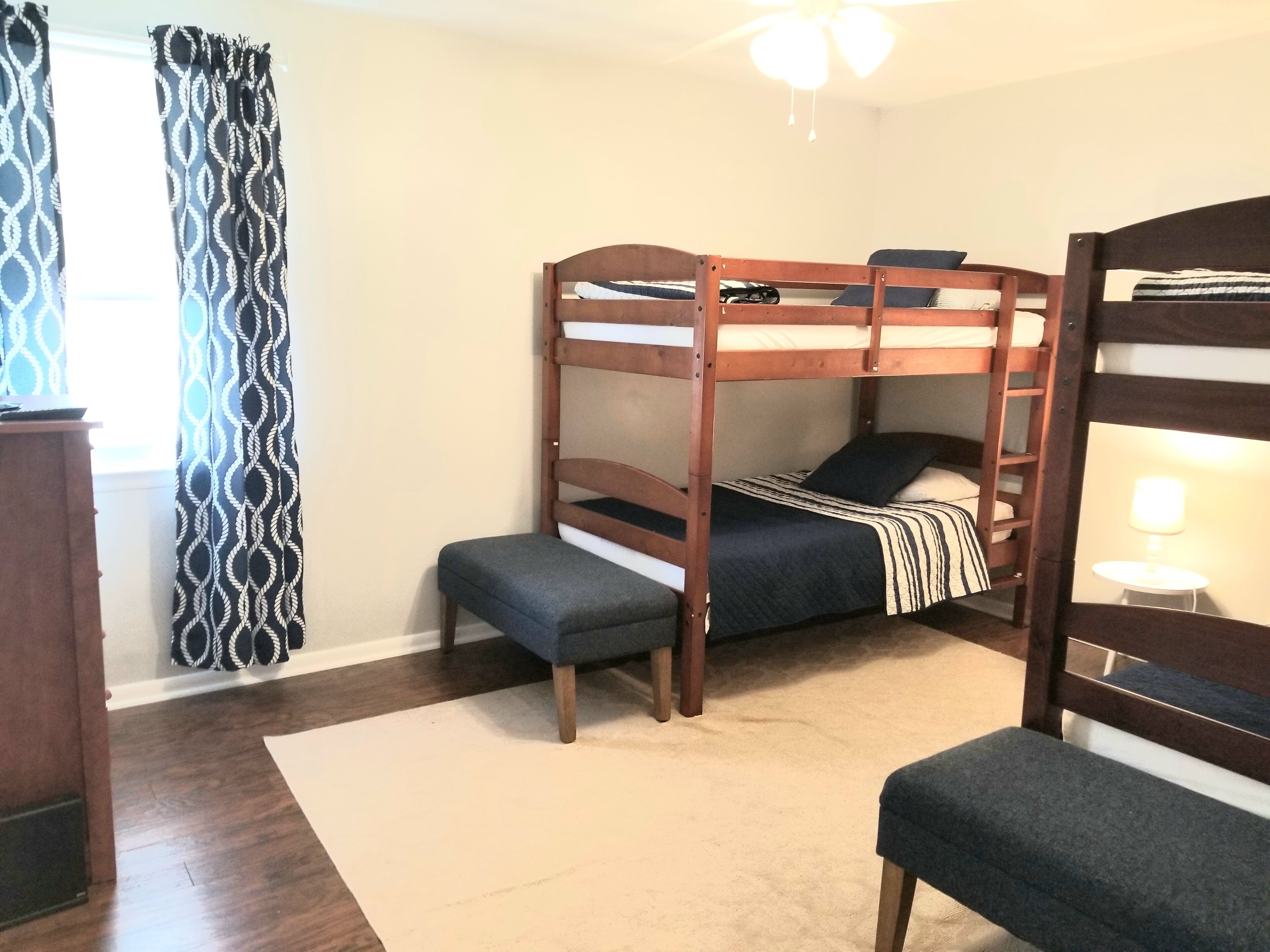 A bunk bed room with a desk and chair, a window with patterned curtains, and a ceiling fan.