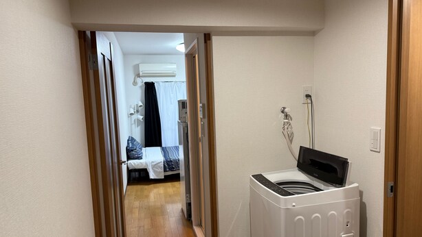 Ein schmaler Flur mit einer Waschmaschine, einem Bett mit gemustertem Überwurf und einer Klimaanlage im Hintergrund.