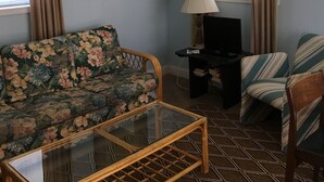 Un salon avec un canapé à motifs floraux, un fauteuil à rayures, une table basse en verre, un meuble pour la télévision avec une lampe, et un tableau accroché au mur.
