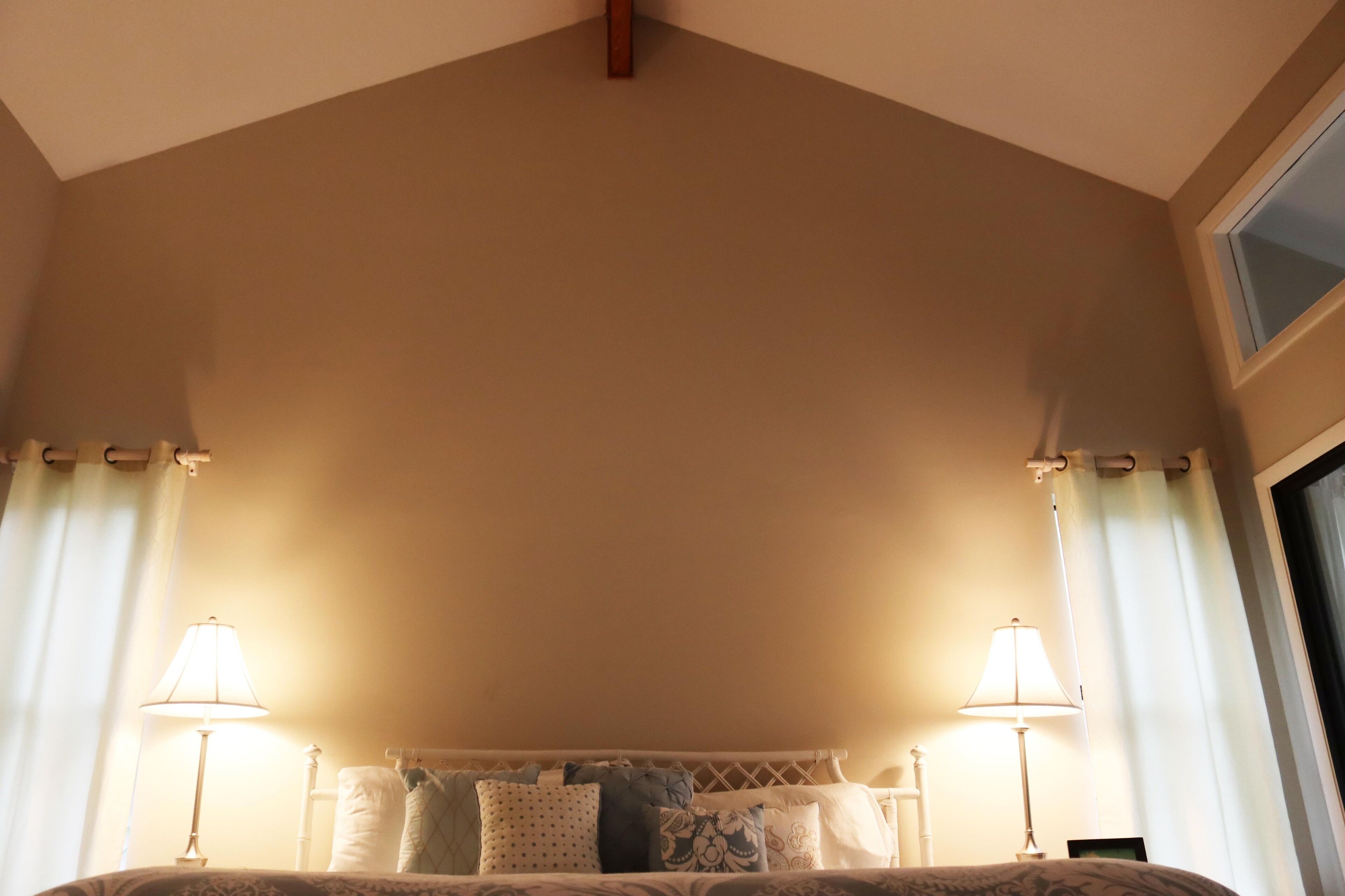 Een slaapkamer met een hoog plafond, een bed met een geborduurd sprei, twee bedlampen en een raam met gordijnen.