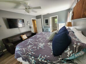 A bedroom with a bed, a ceiling fan, a television, a door, and a small table.