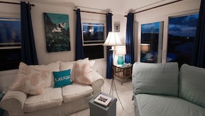 A living room with a sofa, a side table with a lamp, and a view of the outside through large windows. - [A]MOUR HEART OF HOSPITALITY! (Hamilton)