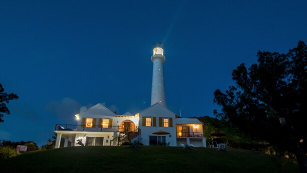 A lighthouse with a white tower and a lit top, surrounded by a house with multiple windows and a balcony. - [A]MOUR HEART OF HOSPITALITY! (Hamilton)