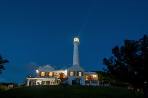 A lighthouse with a white tower and a lit top, surrounded by a house with multiple windows and a balcony.