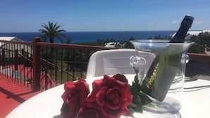 A table with a clear glass pitcher, a bottle, and red roses on a balcony overlooking the ocean. - [A]MOUR HEART OF HOSPITALITY! (Hamilton)